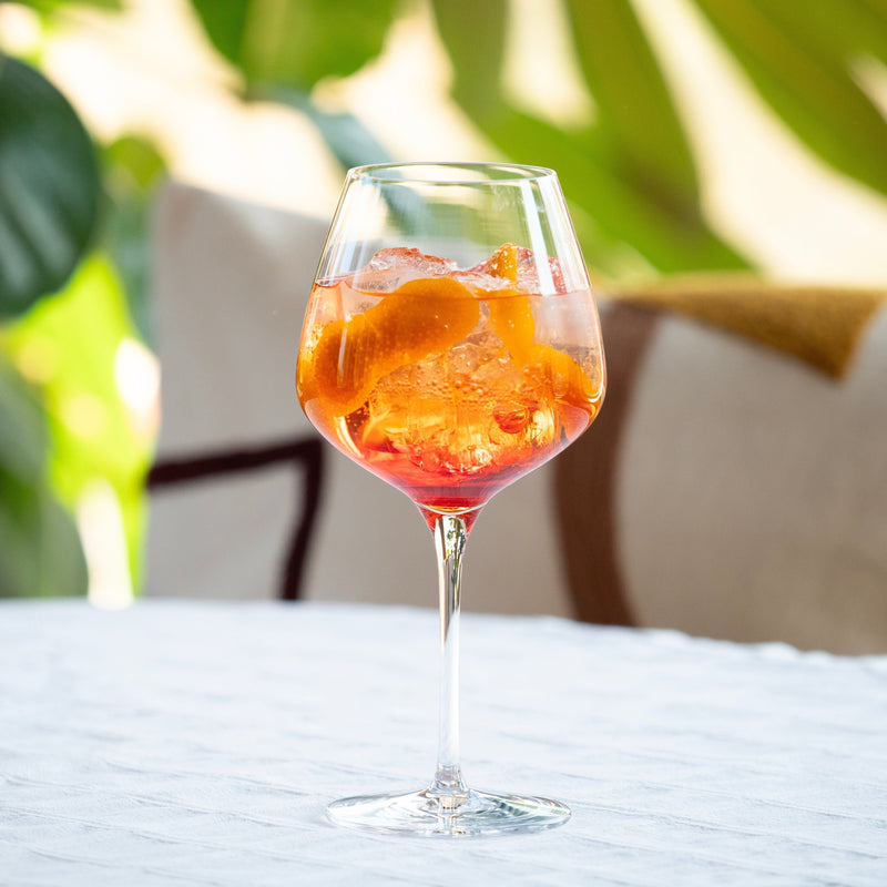 Glass of Aperiniets spritz cocktail recipe with orange twist garnish on a table with green leaves in the background.